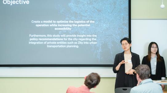 Two students in front of a screen that reads Objective, one speaking with their hands outstretched towards two seated onlookers.