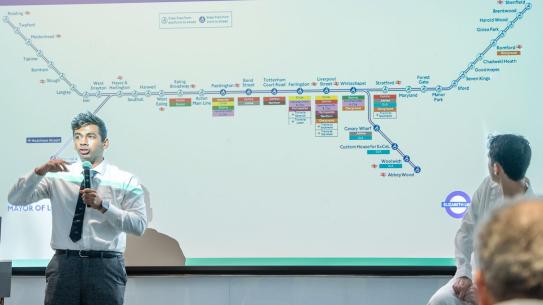 One student in front of a screen showing a London transit map and speaking with his arms outstretched.